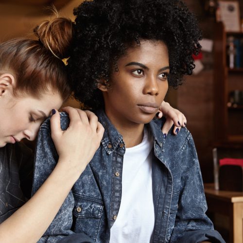 Please forgive me. Mad angry black woman dressed in denim jacket pouting, ignoring apologies of her white redhead female partner who hugging her and begging for forgiveness after having quarrel