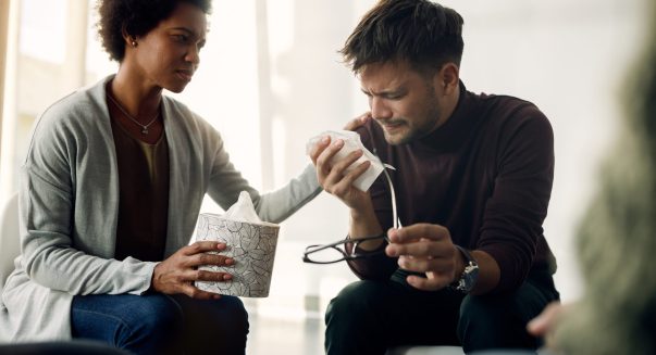 African American woman consoling a crying man who is sharing his issues during group therapy session.